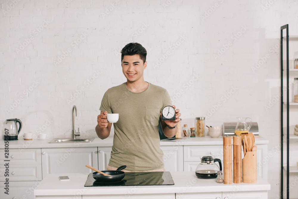 cheerful asian man holding coffee cup and alarm clock while standing near cooking surface with frying pan cheerful asian man holding coffee cup and alarm clock while standing near cooking surface with frying pan