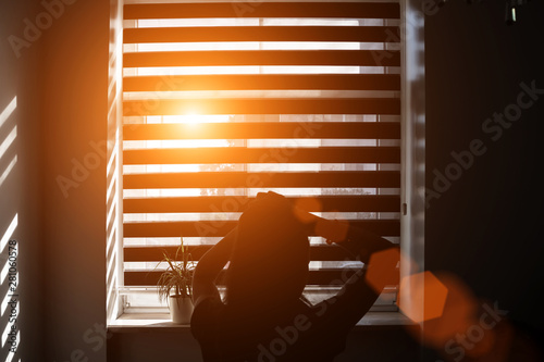 bright sunbeam through gap in blind illuminates silhouette of girl sitting on bed in bedroom and braiding ponytail on her head photo