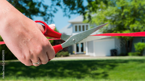 A scissor-cut arm cuts a red ribbon, and a typical American house is visible in the background. Housewarming and buying a home concept