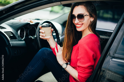 Beautiful young cheerful woman in fashion sunglasses with coffee in hand sitting in black car. Female on road trip drinking coffee inside car. Lifestyle People Travel concepts © victoriazarubina