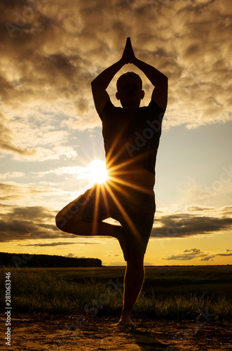 Fototapeta Naklejka Na Ścianę i Meble -  A man in a t-shirt and shorts is practicing yoga in the fresh air. Silhouette against the setting sun and sky. Concept of amateur yoga, healthy lifestyle, meditation practice. Place for text.