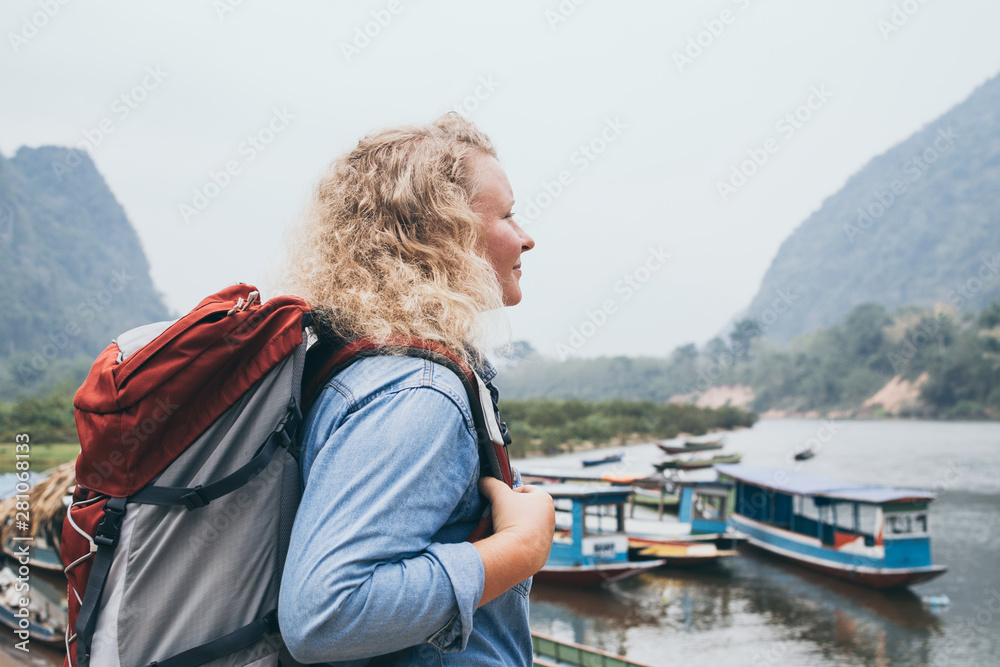 Fototapeta premium Caucasian blonde woman with backpack stands on boat pier in Muang Ngoi village, Laos