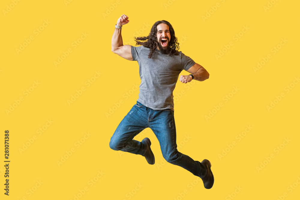 Portrait of happy rejoicing bearded young man with long curly hair in casual grey tshirt jumping and celebrating his vivtory with amazed excited face. indoor studio shot isolated on yellow background.