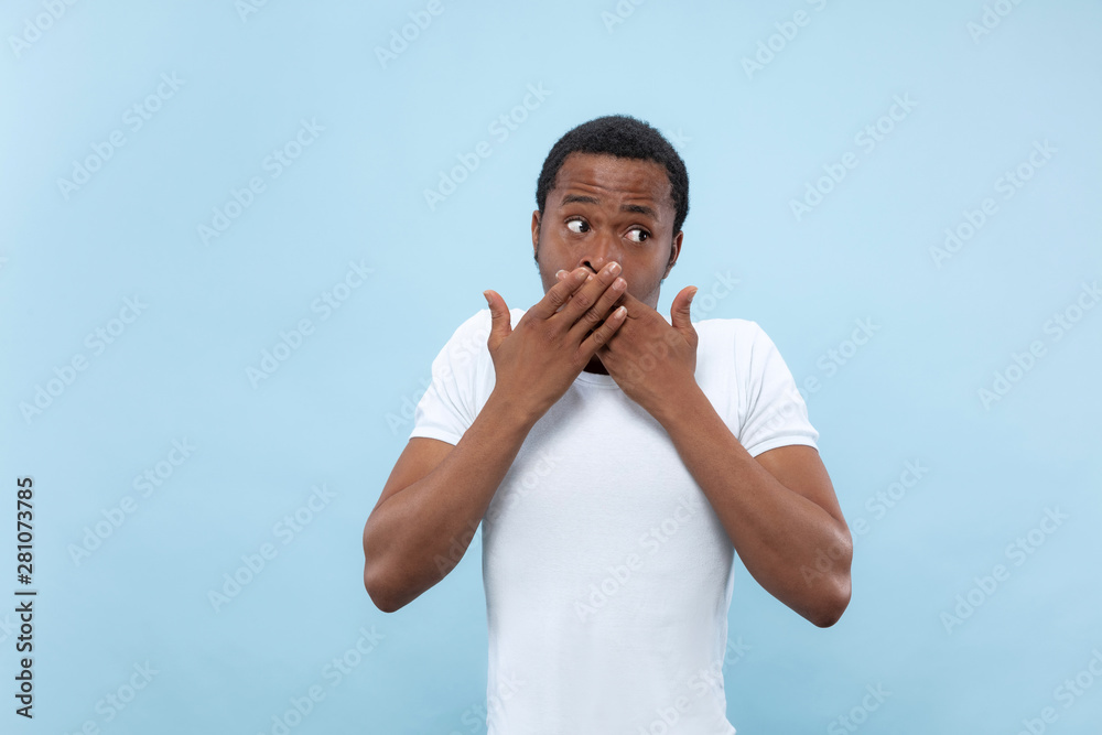 Half-length close up portrait of young african-american man in white shirt on blue background. Human emotions, facial expression, ad concept. Shocked, covering his mouth by his hands. Half-length close up portrait of young african-american man in white shirt on blue background. Human emotions, facial expression, ad concept. Shocked, covering his mouth by his hands.