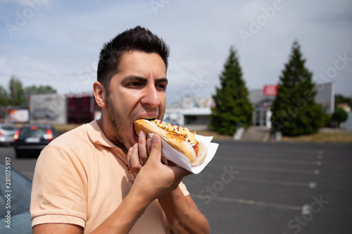 Handsome young brunette man eating hot dog in the parking lot near the gas station.