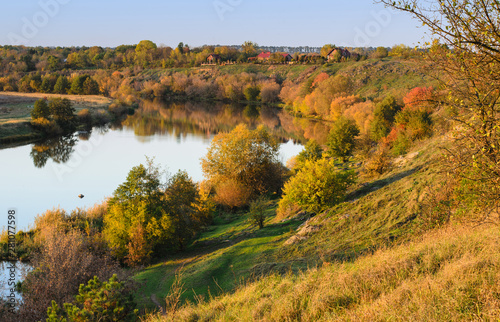Beautiful evening landscape with a river, autumn multicolored trees and small houses on steep banks, October, the Southern Bug river, Central Ukraine