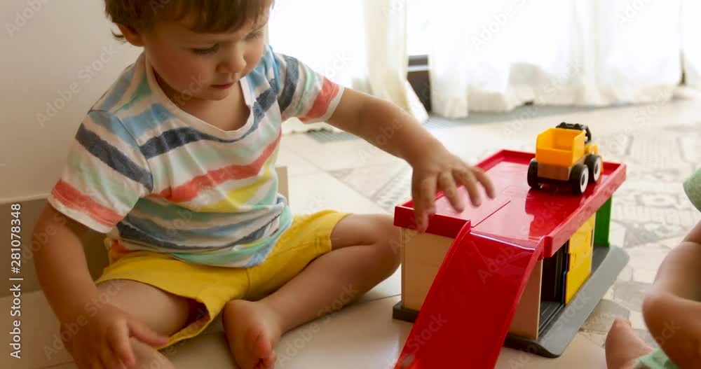 Pretty barefoot toddlers in colorful T-shirts and shorts sitting on floor covered with bright balloons and playing toy car in kindergarten Pretty barefoot toddlers in colorful T-shirts and shorts sitting on floor covered with bright balloons and playing toy car in kindergarten