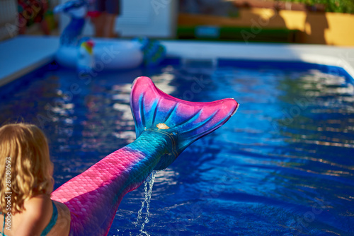  Little girl plays with a mermaid tail in a pool on a sunny day.