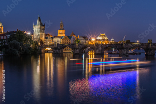 Old Town tower and historic stone bridge with lighting and reflections from the ship on the water. Prague with Charles Bridge at night. Panoramic view over the Vltava