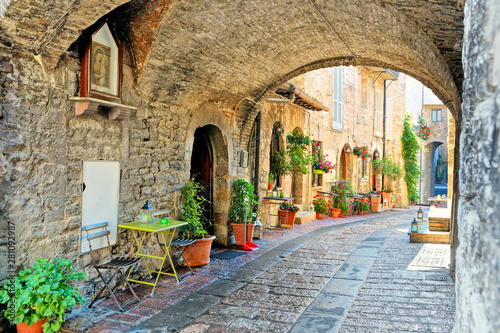 Fototapeta Naklejka Na Ścianę i Meble -  Beautiful arched street in the medieval old town of Assisi with flowers and restaurant tables, Italy