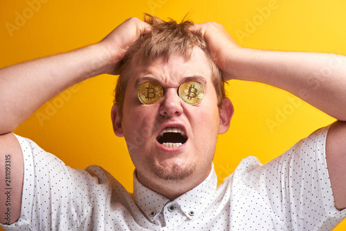 Portrait of upset fat man with bitcoins in eyes. Guy tears hair on head, yells, put money into cryptocurrency and lost everything on yellow background