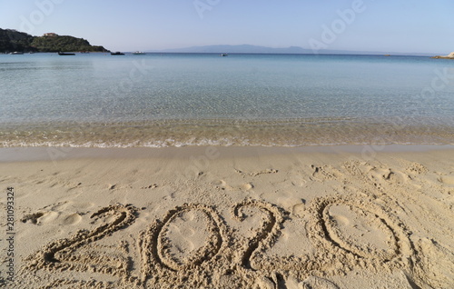 Happy New Year 2020, lettering on the beach with wave and clear blue sea. Numbers 2020 year on the sea shore, message handwritten in the golden sand on beautiful beach background. New Years concept.