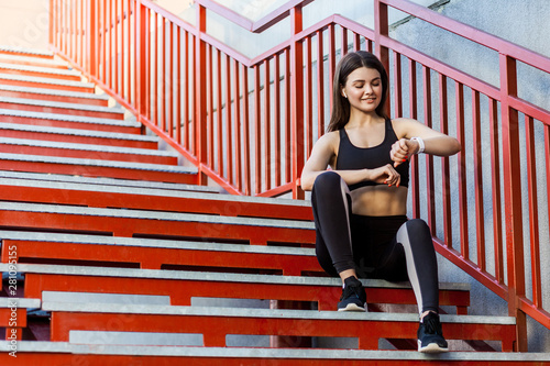 Young sporty beautiful woman having a break sitting on the red stairs after the morning jogging in the modern city  checking result on smartwatch with toothy smile. Outdoor  sport and healthy concept