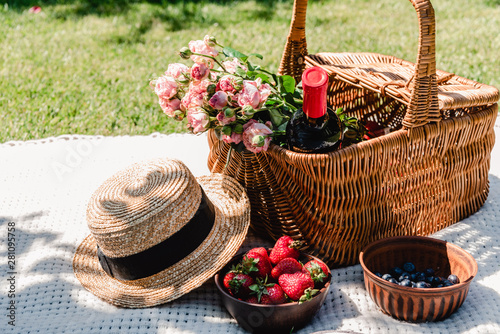 wicker basket with roses and bottle of wine on white blanket near straw hat and berries at sunny day in garden