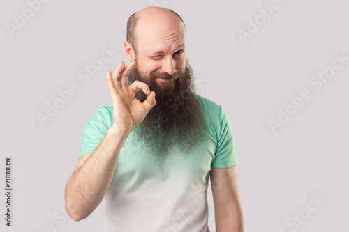 Portrait of funny middle aged bald man with long beard in light green t-shirt standing, winking and looking at camera with Ok sign gesture. indoor studio shot, isolated on grey background.