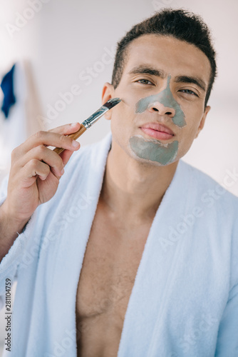handsome man applying clay mask on face with brush while standing in bathrobe