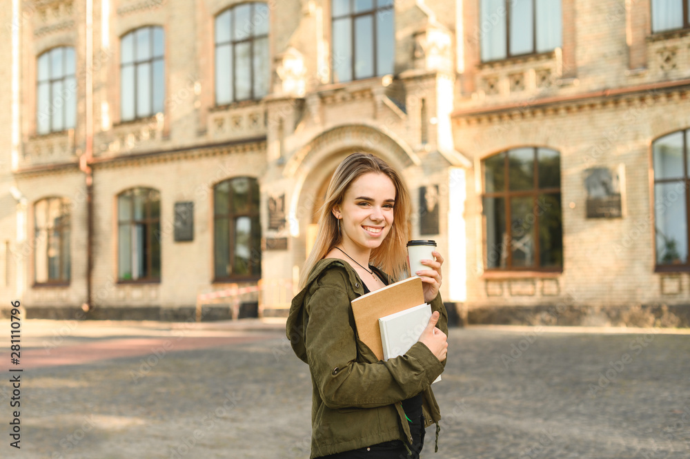 Young talented female student with broad smile in casual clothes standing in front of university building holding books and coffee. Smiling student near college campus with books and take away coffee.