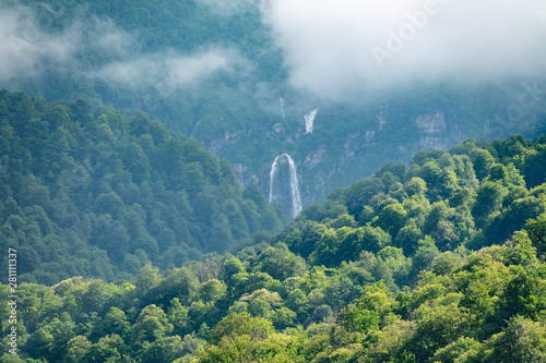 Green mountains with a high waterfall in fog and clouds.
