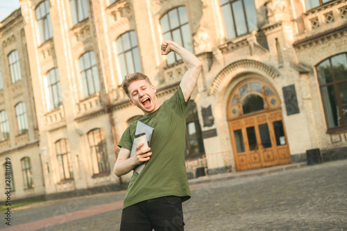Happy cheerful excited male student celebrating his victory with raised hands in front of college campus. The main task completed, exams over, reaching success, winner student with laptop and coffee.