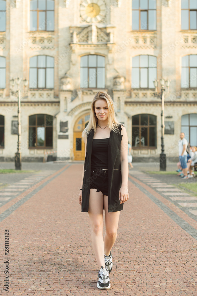 Fototapeta premium Full length portrait of confident attractive female student in total black look walking near the college campus. Succesful stylish student girl posing near the university building. Vertical shot.
