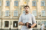 Portrait of a young handsome bearded man in striped shirt holding professional camera, shooting outdoors, stylishly dressed, photographer, close up, brutal, street photo, architecture background.