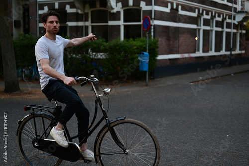 Fototapeta Naklejka Na Ścianę i Meble -  Dutch male student pointing to the left while riding on his bicycle through an old Dutch street in Amsterdam, the Netherlands