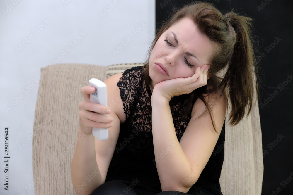 Portrait of a pretty beautiful fashionable adult brunette girl in a black dress. Sits on the sofa with the remote control directly in front of the camera, showing different poses and emotions Portrait of a pretty beautiful fashionable adult brunette girl in a black dress. Sits on the sofa with the remote control directly in front of the camera, showing different poses and emotions