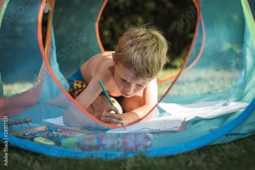 Preschool boy draws something in a tent in the garden during summer day.