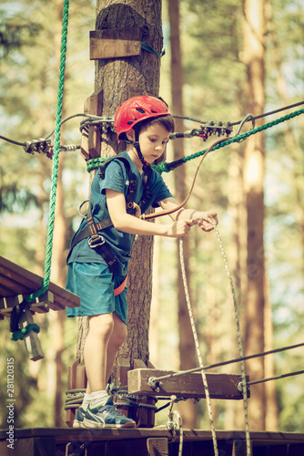 Child in forest adventure park. Kid in red helmet and blue t shirt climbs on high rope trail. Agility skills and climbing outdoor amusement center for children. Young boy plays outdoors.