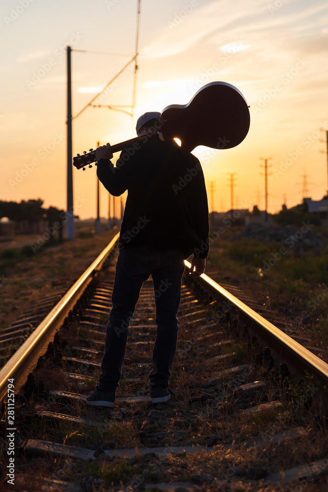 A young guy stands on the rails with a guitar on his shoulders at sunset