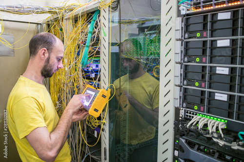 The man works in the server room of the datacenter. A technician measures the signal level in a fiber optic cable. The system administrator is testing the network with a reflectometer. photo