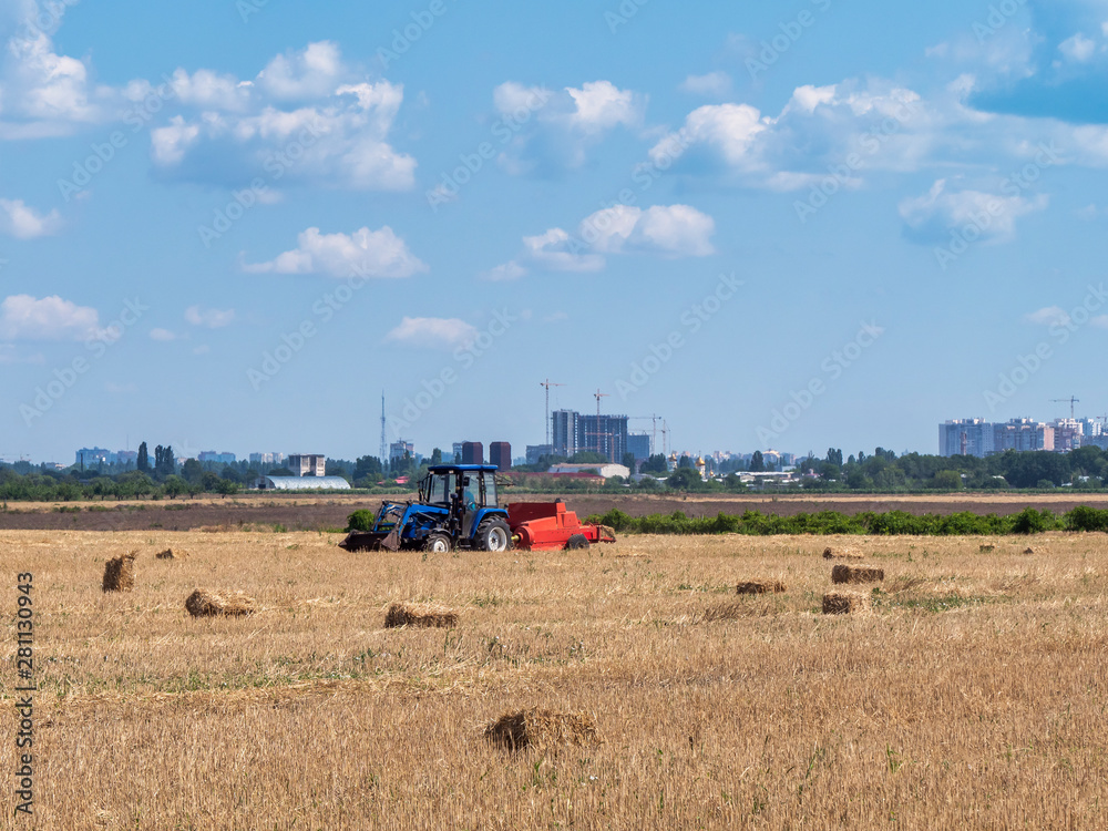 Fototapeta premium Hay is packaged in briquettes on a field under a blue sky with clouds. Works special agricultural equipment. The village and the city . In the background is the construction of the city. Urbanization.