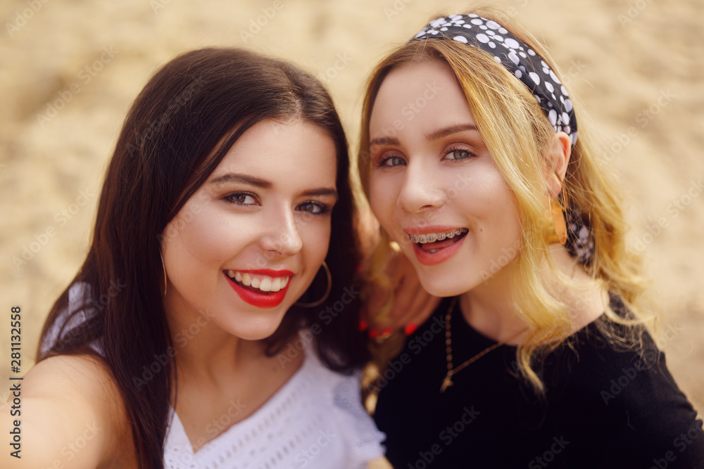 Fototapeta premium Girls on the beach stand on the background of sand. The brunette in white and the blonde with a bandana on her head. Beautiful girls look at the camera and take a selfie. Girls make photos on camera.
