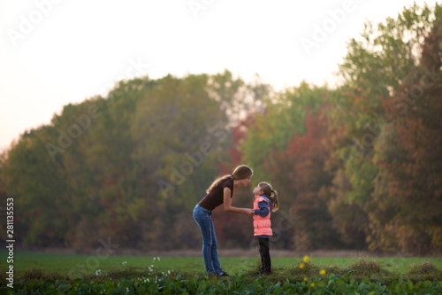 Profile full-length portrait of young slim attractive mother talking to child girl standing in green meadow holding hands outdoors on forest trees blurred background.