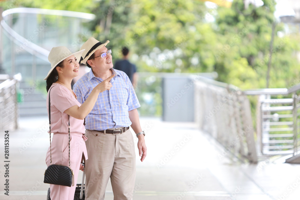 asian traveler couple husband and wife in pink and blue shirt with white nice hat and luggage walking in downtown, she pointing something with right copy space