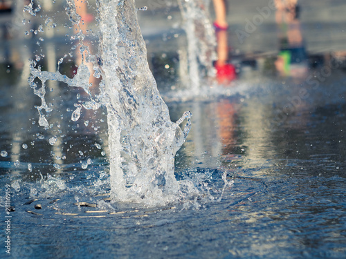 Close up view on water streams flowing out of stainless grate of dry fountain. Kids bare feet cooling near fountain. Town square in summer. Selective soft focus. Blurred background