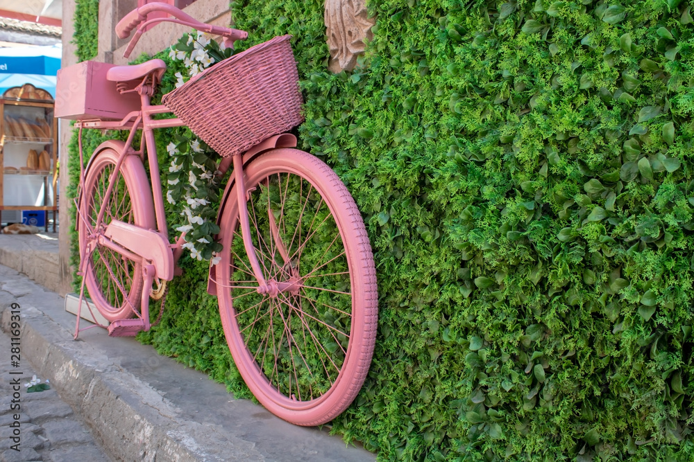 Old pink basket bike standing on pavement. It's attached to the wall.