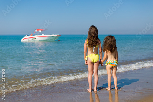 little girls in swimsuits on the beach standing back and looking at the boat in the sea Children on vacation
