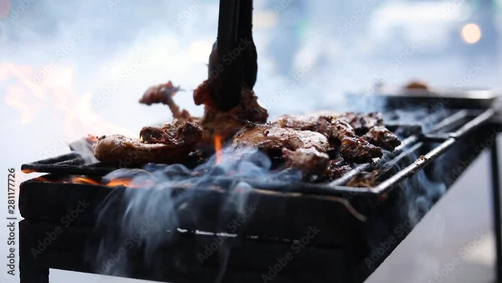 Slow Motion shot of an Asian street vendor cooking Ayam Bakar (Grilled Chicken) on a smoky grill. The cook is flipping some of the pieces of chicken.