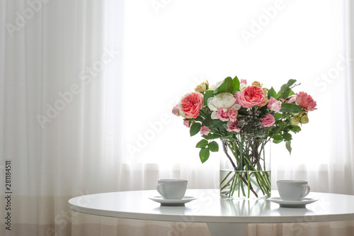 Vase with beautiful flower bouquet and cups of coffee on table in room. Space for text