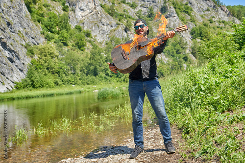 A musician plays a burning acoustic guitar against the backdrop of a natural landscape