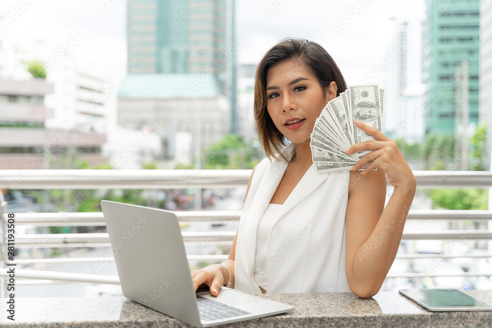 Beautiful girl smiling in business woman clothes using laptop computer and show money US dollar bills in hand , urban city background Beautiful girl smiling in business woman clothes using laptop computer and show money US dollar bills in hand , urban city background
