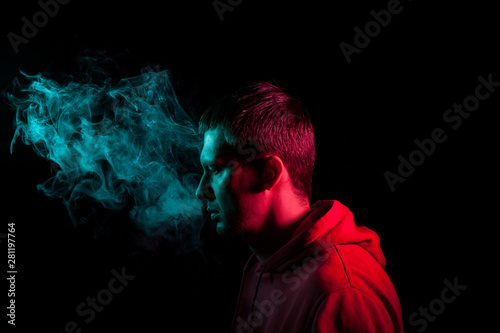 Close up portrait of the face of an adult serious man exhales green toxic smoke while smoking e-cigarette and vape illuminated with blue and red colored light on a black background. Harm to health. © Aleksandr Kondratov