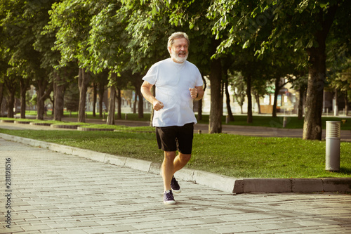 Senior man as runner with armband or fitness tracker at the city's street. Caucasian male model practicing jogging and cardio trainings in summer's morning. Healthy lifestyle, sport, activity concept.