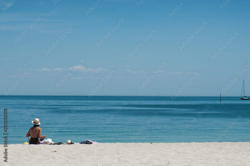 Portrait of woman in swimsuit and hat sitting on the beach on back view with sailboat on background Portrait of woman in swimsuit and hat sitting on the beach on back view with sailboat on background
