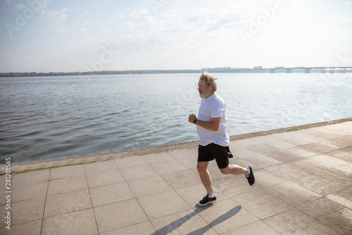 Senior man as runner with armband or fitness tracker at the riverside. Caucasian male model practicing jogging and cardio trainings in summer's morning. Healthy lifestyle, sport, activity concept.