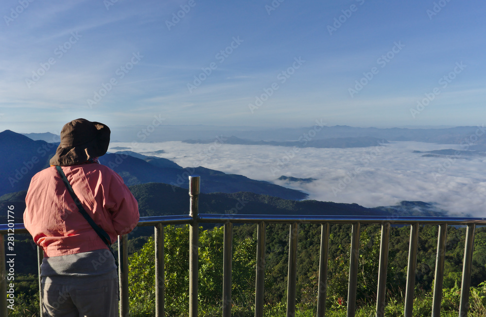 Female asian traveller wearing pink jacket and brown hat holding stainless steel fence looking to the scenic view of fog, mist, forest and mountain at Doi inthanon Chiangmai Thailand on sunny day