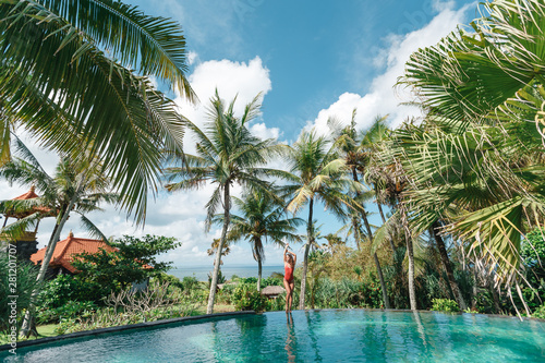 Girl in red swimsuit on the edge of pool in the jungles with ocean on the background. Palms around and crystal clean water. Luxury resort on Bali island