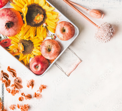 Tray with apples, sunflowers and pomegranate on white table with autumn leaves and botanical decoration, top view. Flat lay. Modern. Minimal. Fall seasonal layout. Copy space for your design.