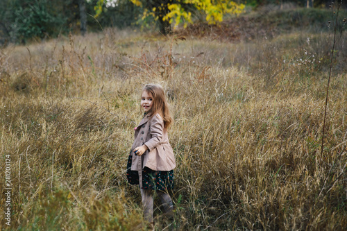fair-haired girl in a beige coat on the dry grass and autumn trees background.